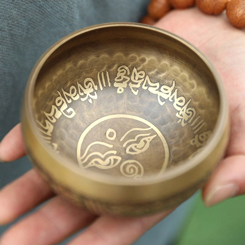 Buddhist singing bowl held with prayer beads, displaying hammered brass with golden inner symbols and sacred script