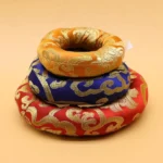 Stacked handcrafted Buddhist singing bowl cushions with golden brocade embroidery in multiple colors including red, navy, and gold - top view