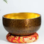 Handcrafted copper singing bowl with golden interior displayed on red embroidered meditation cushion, front view