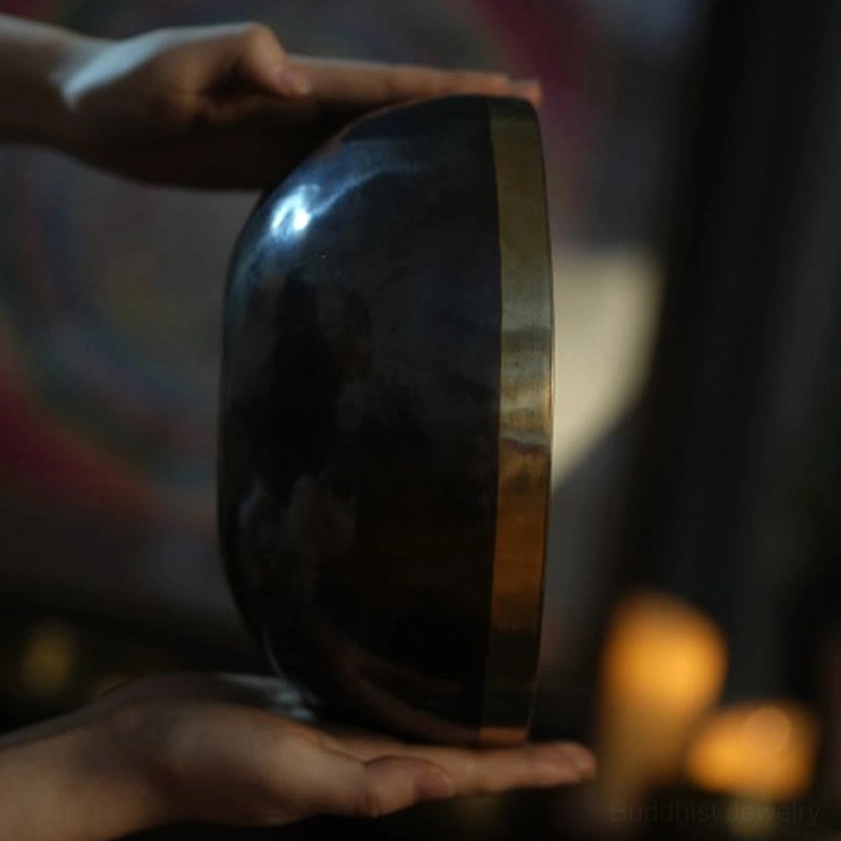 Buddhist singing bowl held in two hands against candlelit background, demonstrating handheld playing technique for meditation sound bath