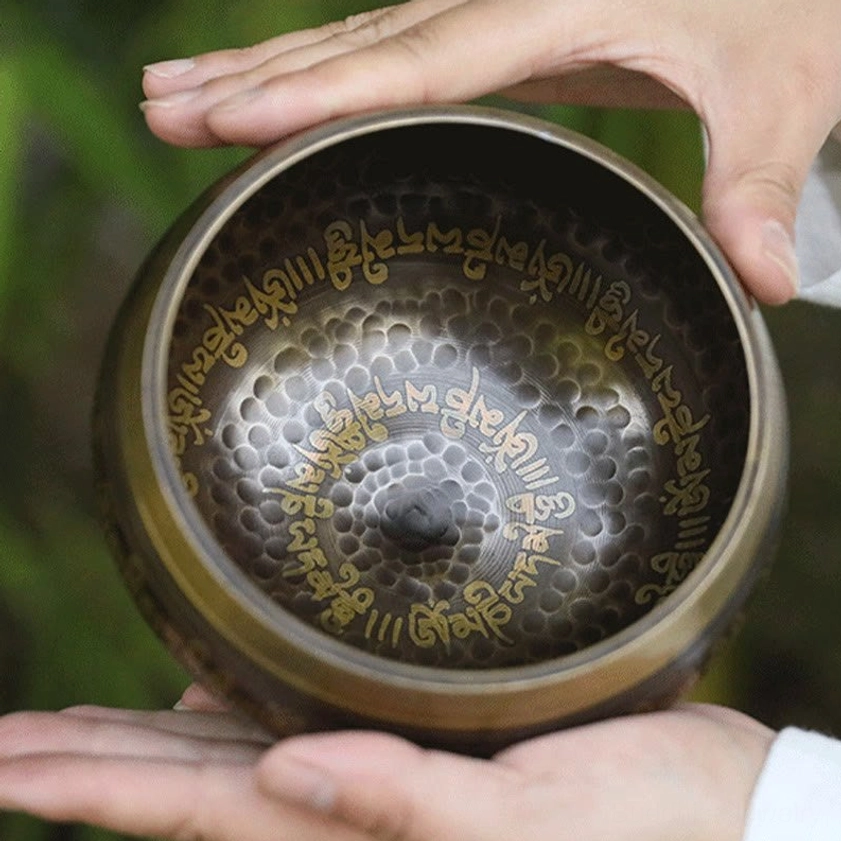 Interior view of handcrafted Buddhist singing bowl showing hammered dotted texture pattern and golden Sanskrit inscriptions on gilded rim, held in hand with bamboo forest backdrop