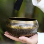 Handcrafted Buddhist singing bowl held in two hands, displaying bronze finish with golden Sanskrit mantras, hammered texture, and gilded rim, photographed in serene bamboo forest setting