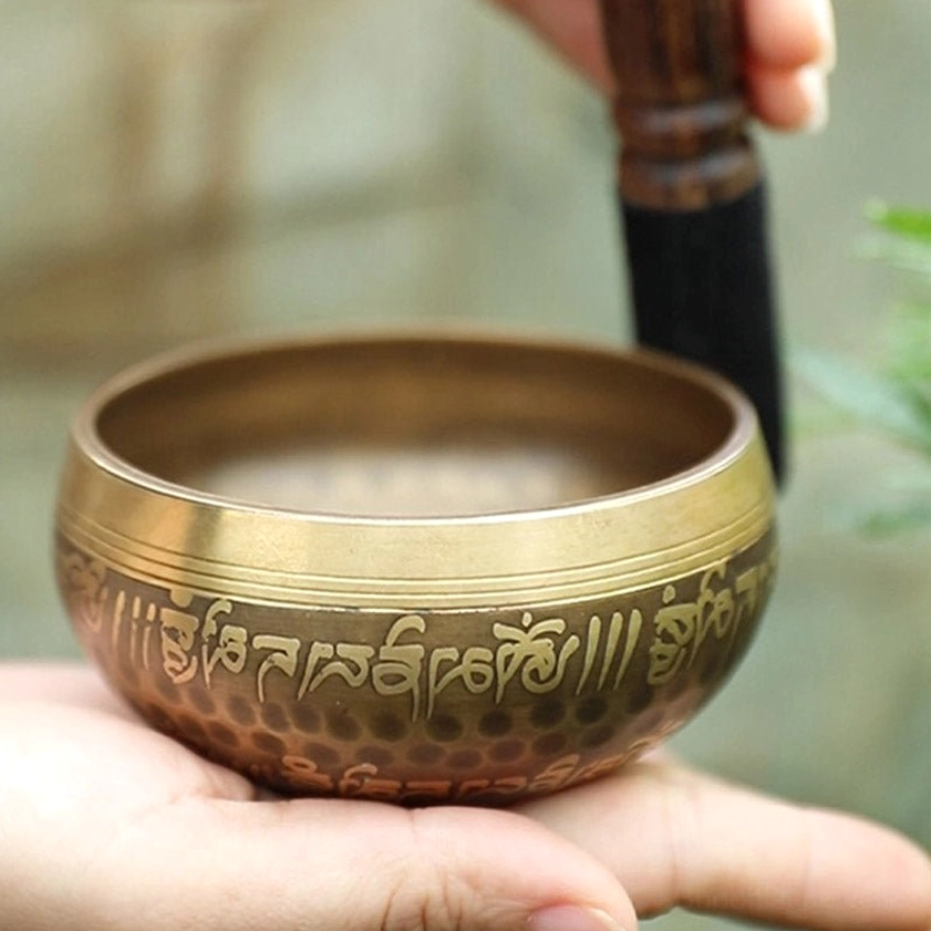 Antique bronze Buddhist singing bowl held in hands, side profile view displaying engraved mantras and hammered copper body