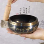 Handcrafted Buddhist singing bowl with golden Om Mani Padme Hum mantra engraving, shown in side handheld view with wooden mallet against calligraphy background