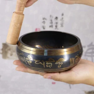 Handcrafted Buddhist singing bowl with golden Om Mani Padme Hum mantra engraving, shown in side handheld view with wooden mallet against calligraphy background