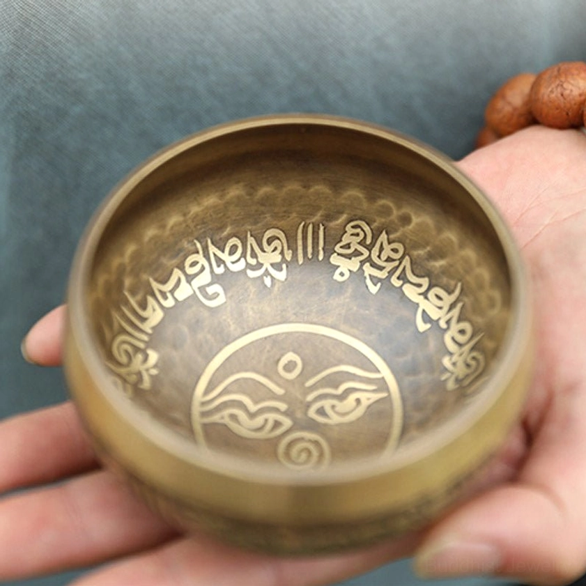 Buddhist singing bowl held with prayer beads, displaying inner eye symbol and rim engraved sacred mantras