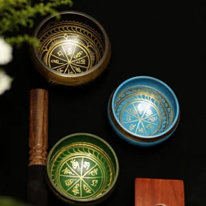 Handcrafted Buddhist singing bowl set with multiple colored brass bowls, gold Sanskrit engravings, wooden striker, and embroidered silk cushions displayed in flat lay composition