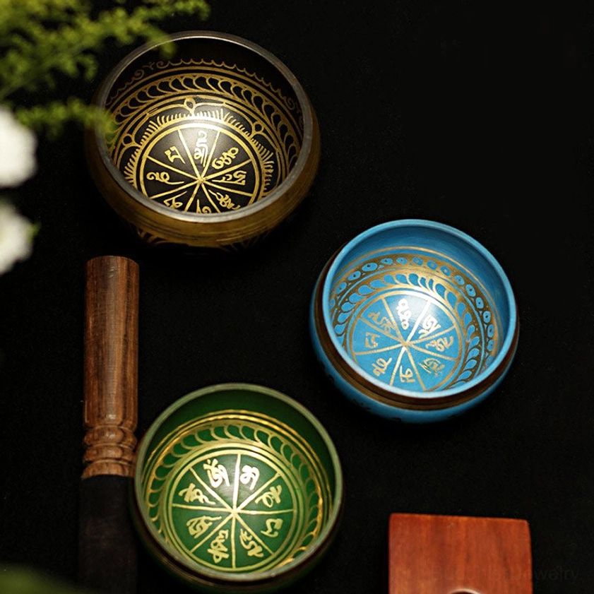 Handcrafted Buddhist singing bowl set with multiple colored brass bowls, gold Sanskrit engravings, wooden striker, and embroidered silk cushions displayed in flat lay composition