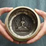 Top view of handcrafted Buddhist singing bowl held in hands showing engraved Buddha statue and sacred scriptures in bronze finish