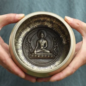 Top view of handcrafted Buddhist singing bowl held in hands showing engraved Buddha statue and sacred scriptures in bronze finish