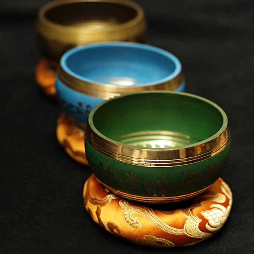 Three Buddhist meditation singing bowls in blue, green, and brass with gold decorative rims resting on embroidered silk cushions from angled perspective