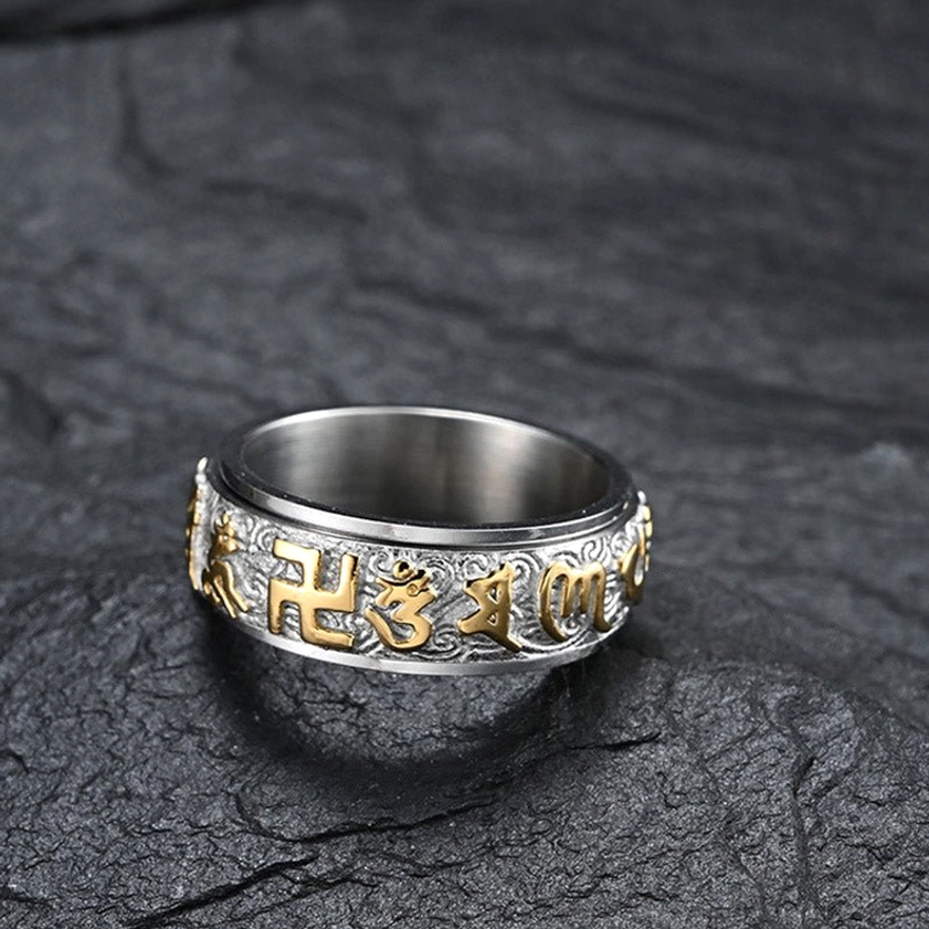 Buddhist mantra spinner ring with rotating band, silver and gold detailing, swastika and Sanskrit engravings clearly visible, front view on stone texture