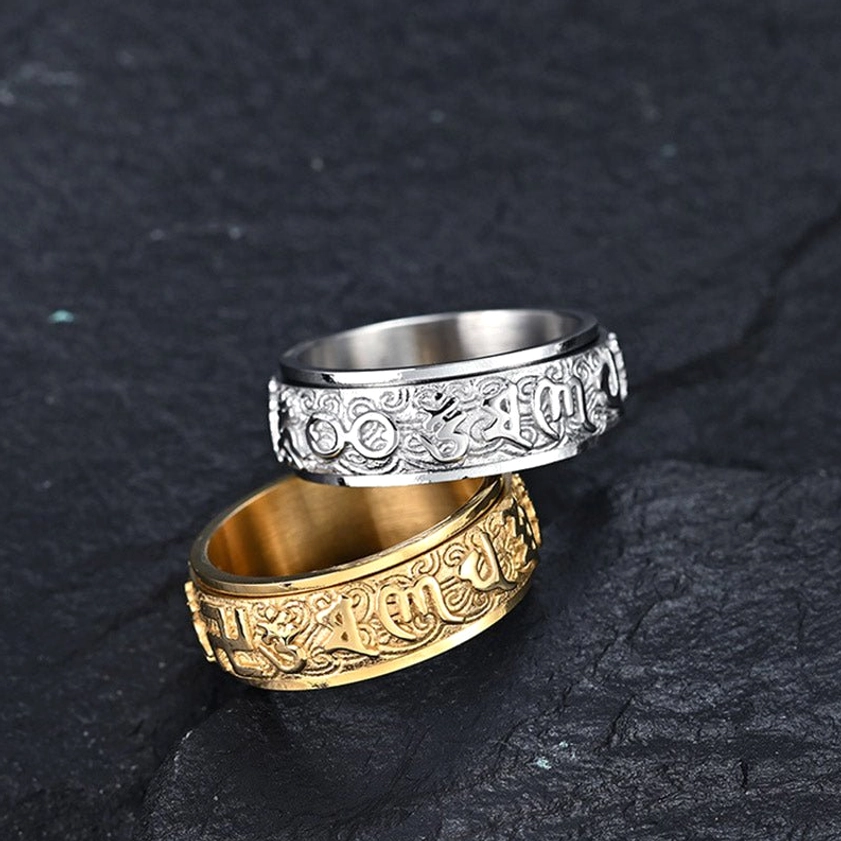Two stacked Buddhist mantra spinner rings in silver and gold tones with engraved Sanskrit patterns, top-down view on dark slate background