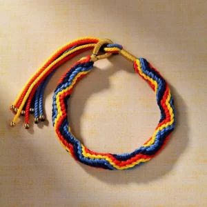 Handwoven multicolor Buddhist string bracelet with chevron braid pattern and gold metal tassel accents, overhead side view showing vibrant red blue yellow and green threads