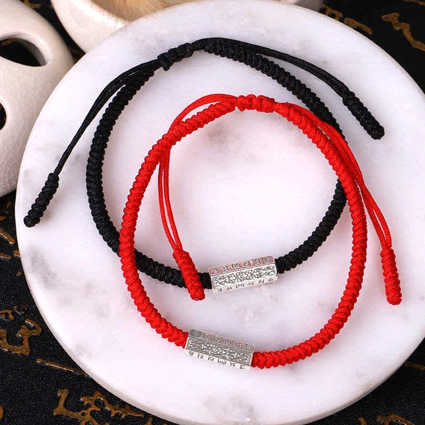Red and black Om Mani Padme Hum Buddhist string bracelets with engraved silver metal beads on braided rope, arranged on marble plate with vintage calligraphy backdrop