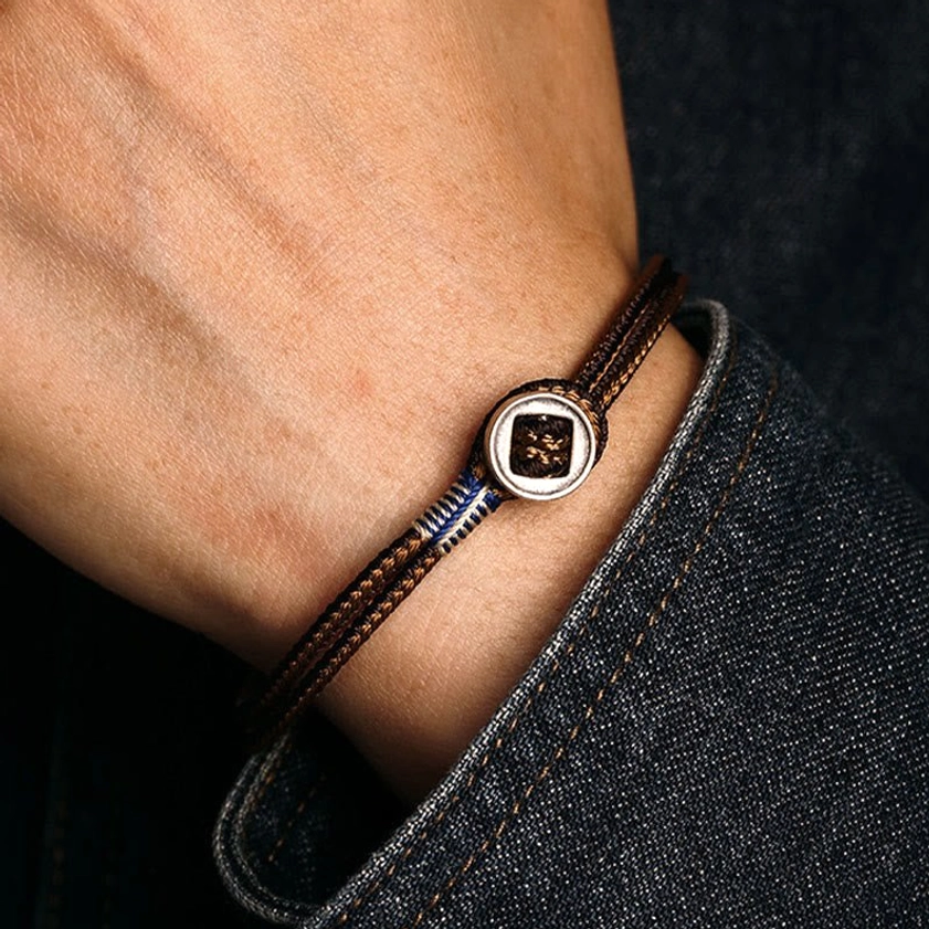 Brown woven Buddhist protection bracelet worn on left wrist over denim fabric, on-wrist product demonstration