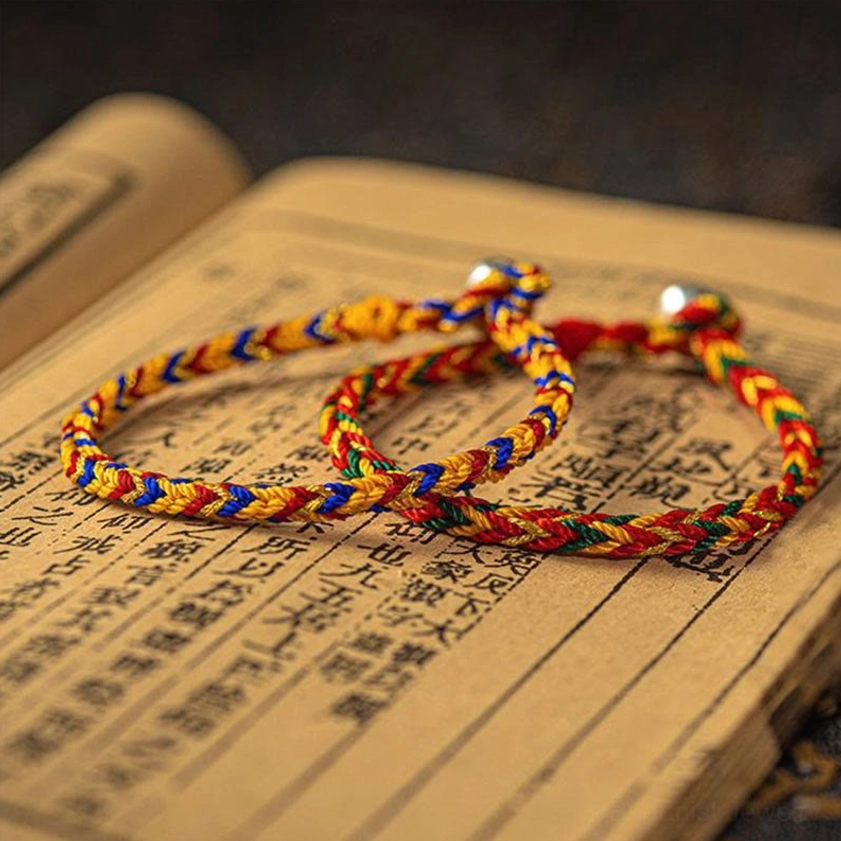 Two multicolored lucky Buddhist protection bracelets displayed on ancient calligraphy page, showing detailed twisted weave and metal bead clasps