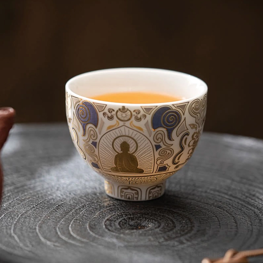 Filled Buddhist ceramic tea cup with golden yellow tea, eye-level front view showcasing gilded Buddha artwork and matching decorative designs on carved wooden tray