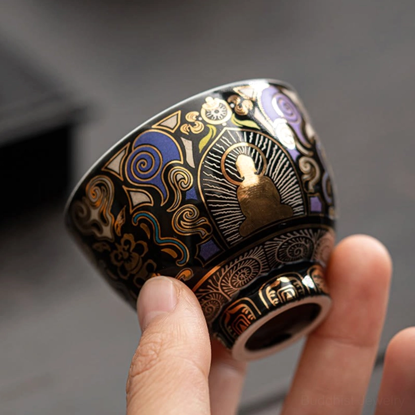 Bottom view of black ceramic Buddhist tea cup showing layered detailed patterns and cup foot, held against light gray background