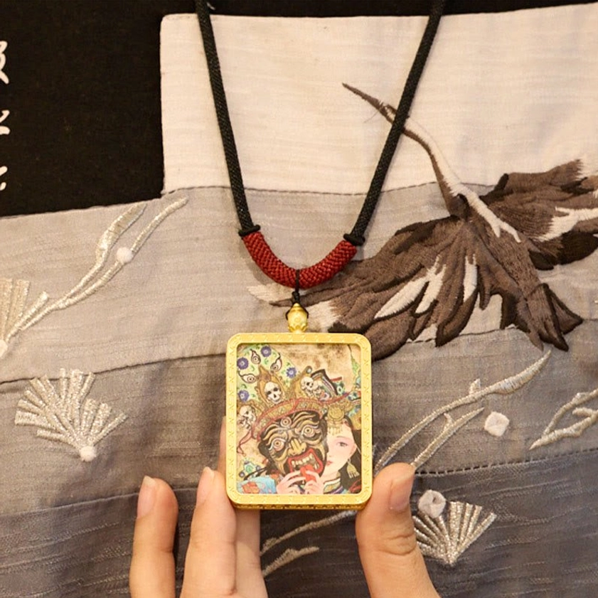 Buddhist Thangka pendant held against embroidered fabric showing intricate gold frame and sacred artwork