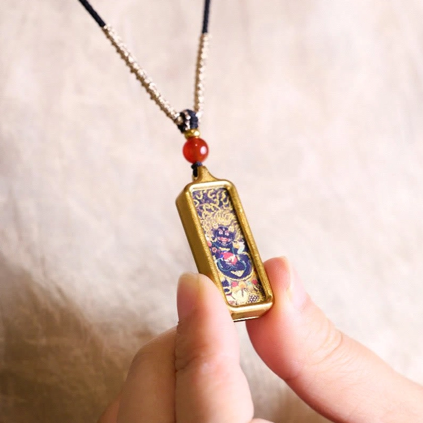 Buddhist thangka pendant necklace held between fingers, showing twisted silver cord, red agate bead and wrathful deity thangka artwork detail
