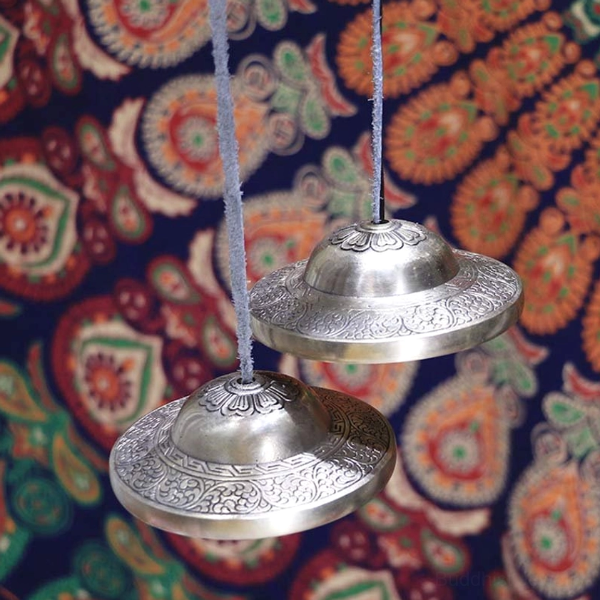 Front hanging view of authentic Buddhist tingsha cymbals showing silver-bronze tone with embossed decorative textures and ethnic fabric background