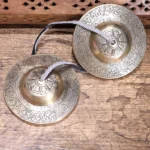 Pair of handcrafted Buddhist copper tingsha bells with embossed floral motifs and leather connecting straps, displayed on weathered wooden surface