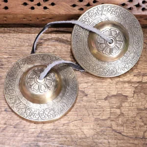 Pair of handcrafted Buddhist copper tingsha bells with embossed floral motifs and leather connecting straps, displayed on weathered wooden surface
