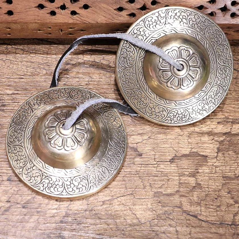 Pair of handcrafted Buddhist copper tingsha bells with embossed floral motifs and leather connecting straps, displayed on weathered wooden surface