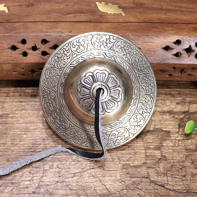 Individual Buddhist tingsha bell featuring intricate floral engravings and black leather hanging strap on carved wooden backdrop