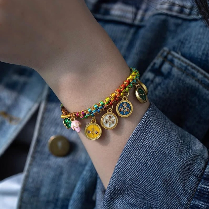 Five Gods of Wealth bracelet worn on wrist over denim clothing, displaying braided Buddhist string with hanging metal wealth deity charms