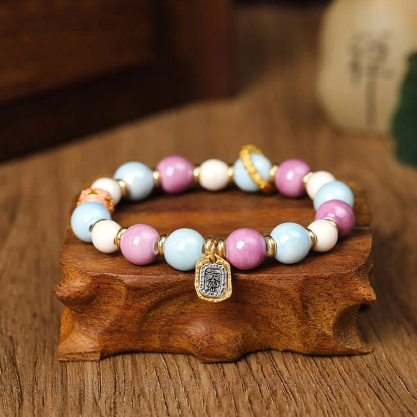 Buddhist Zakiram Goddess of Wealth bracelet with rectangular gold charm, pastel porcelain beads, and metal accents on wooden display