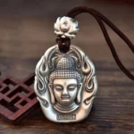 Matte silver Tathagata Buddha head pendant necklace with lotus crown, flame pattern border, and brown braided cord on wooden desk with Chinese carved decor