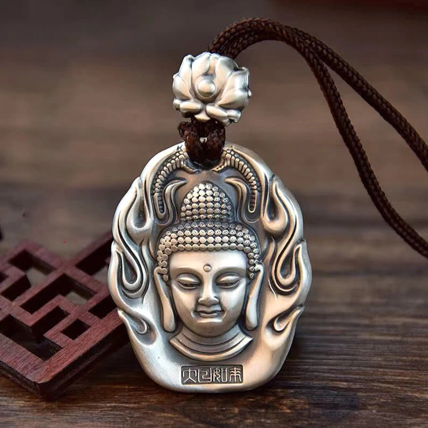 Matte silver Tathagata Buddha head pendant necklace with lotus crown, flame pattern border, and brown braided cord on wooden desk with Chinese carved decor