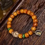 Handcrafted camel bone Buddha head bracelet flat lay featuring amber barrel beads, green jade spacers, carved Buddha charm, and metal protective pendant on dark wood surface