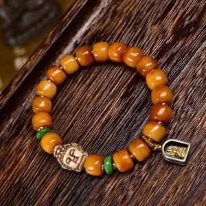 Handcrafted camel bone Buddha head bracelet flat lay featuring amber barrel beads, green jade spacers, carved Buddha charm, and metal protective pendant on dark wood surface