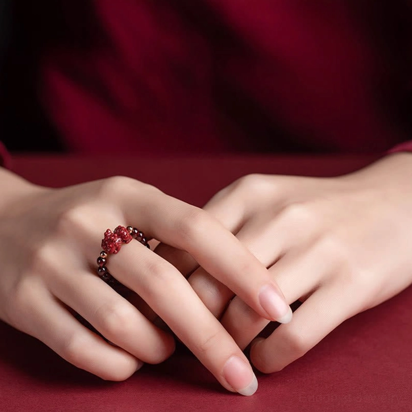 Hand wearing carved cinnabar PiXiu Buddhist ring on middle finger with dark beaded band against wine-red tabletop, showing wealth-protection charm detail