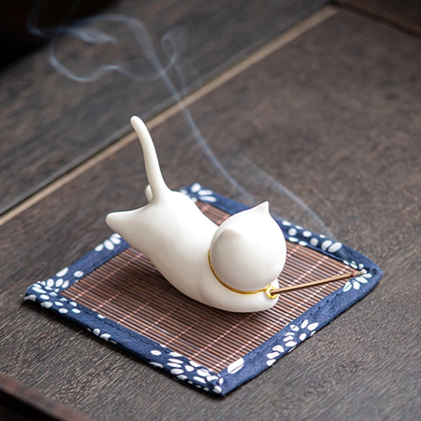 Cat ceramic incense burner from right oblique angle with inserted incense stick and visible smoke trails on dark wooden surface