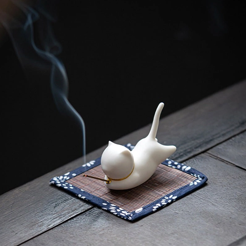 Cat incense burner from left side view with thin curling incense smoke and floral trim mat on grey tiled tabletop
