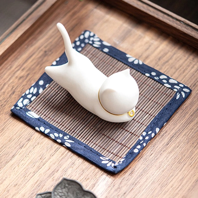 Top-down view of smooth white cat ceramic incense burner with inserted incense stick on floral trim bamboo mat, wooden tray