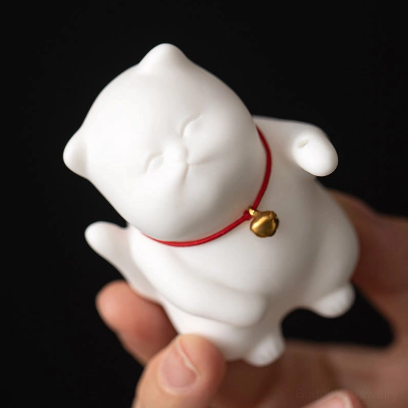 Extreme close-up of white ceramic cat with red collar and gold bell held in fingers, showcasing matte finish and peaceful expression details