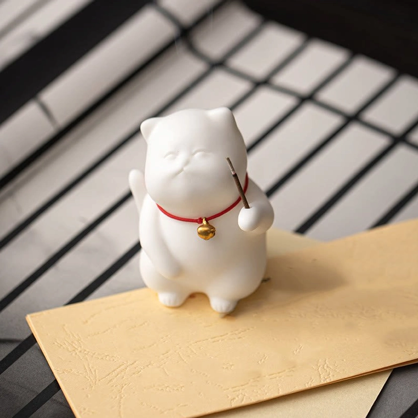 Front perspective view of white ceramic cat incense burner with red collar holding incense stick on desk with grid pattern backdrop