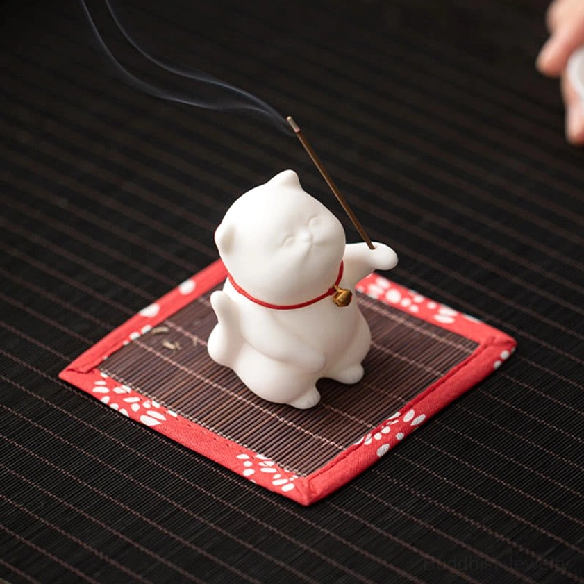 Chubby white porcelain cat incense holder with red collar and gold bell from above, lit incense emitting smoke