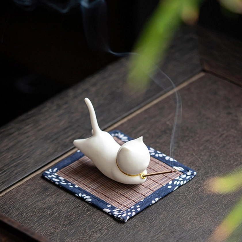 Cat-shaped ceramic incense holder from low side angle showing subtle rising incense smoke and gold neck decoration on dark wooden desk