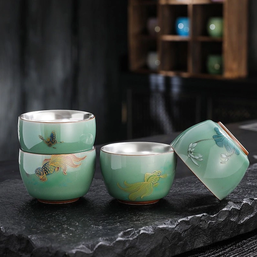 Collection of handcrafted celadon Buddhist tea cups featuring butterfly, lotus and koi fish designs with silver-lined interiors displayed on dark slate and wooden shelf