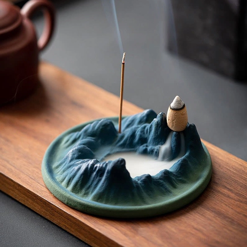 Buddhist ceramic backflow incense burner with mountains and river design on wooden tea table, showing matte ceramic finish with teal base and indigo mountain surface