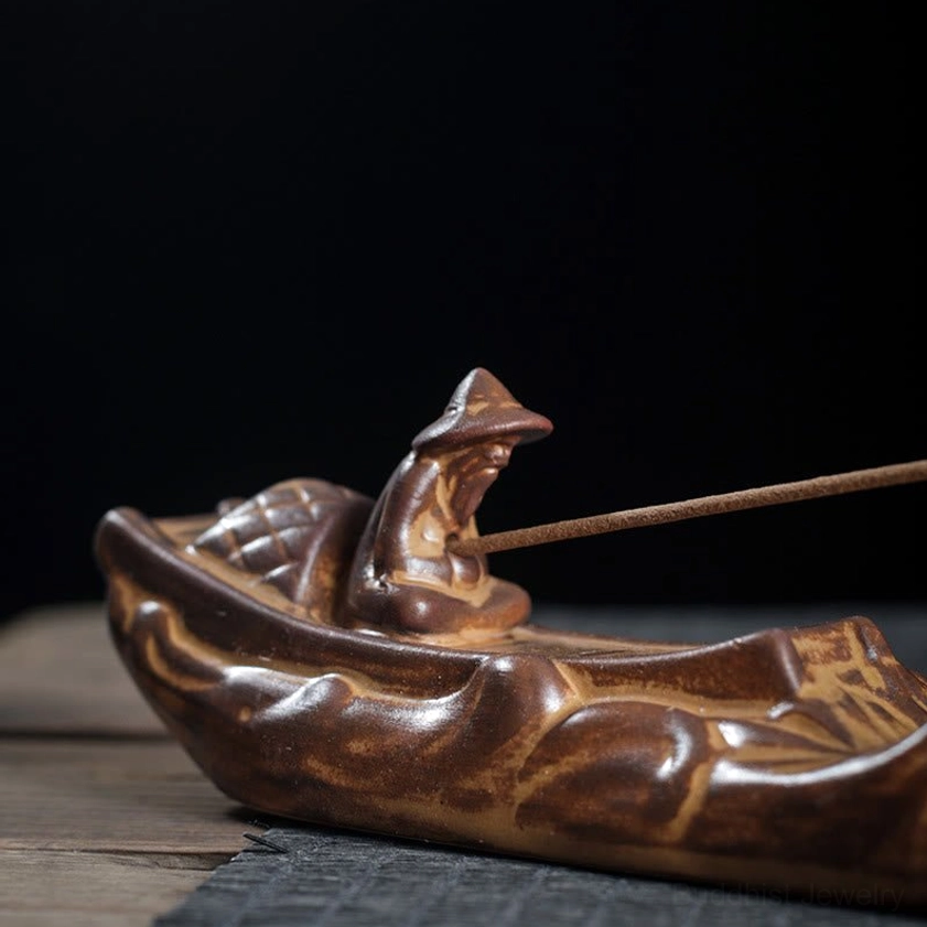 Close-up side view of handcrafted boat incense burner with carved fisherman figure holding incense stick on wooden desktop