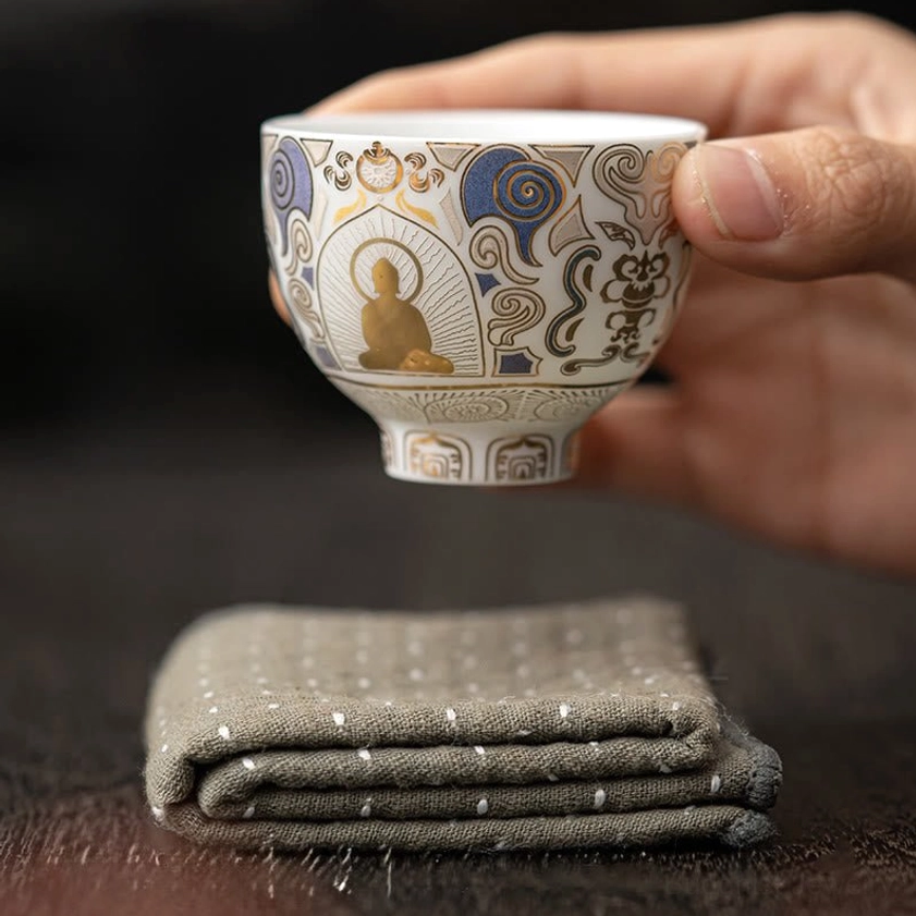 Side view of authentic handmade ceramic kung fu teacup with Buddha motif and gold detailing, held by hand on wooden tabletop with decorative linen beneath