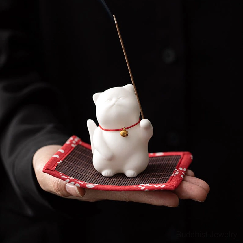 Overhead close-up of small white ceramic cat incense burner resting in human palm with floral fabric coaster, demonstrating compact portable design