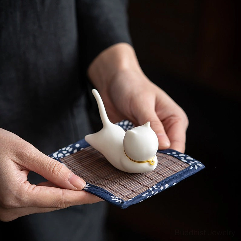 Hand-held ceramic cat incense burner at eye level with blue and white floral edge bamboo mat, studio indoor background